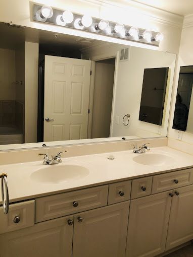 Bathroom with double sinks, large mirror, white cabinets, and overhead lights.