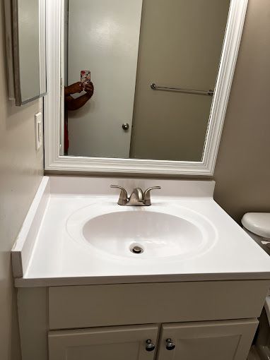 White bathroom vanity with oval sink, faucet, and mirror. A person holding a phone is reflected in the mirror.