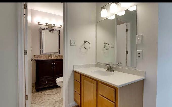 Bathroom interior: two vanities, one wooden and one with light wood cabinets, mirrors, and white walls.