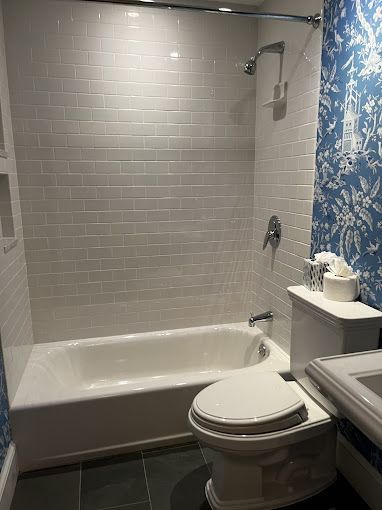 Bathroom with white subway tile shower, white tub, toilet, and blue patterned wallpaper.