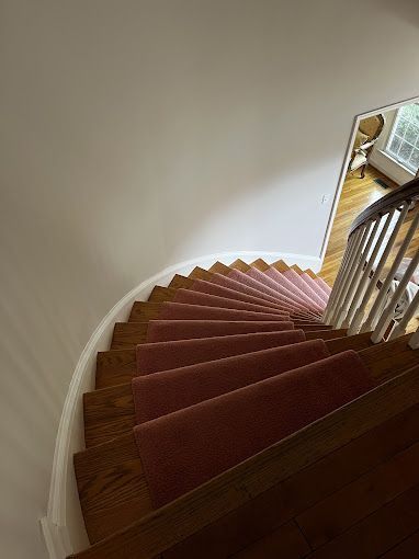 Curving staircase with pink carpet runner and wooden steps, leading towards a bright window.