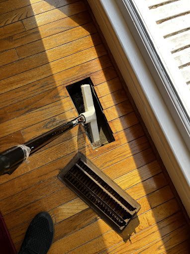 Vacuuming a floor vent with a vacuum cleaner on a hardwood floor, next to a window.