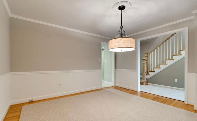 Empty dining room with beige walls, white wainscoting, and a light fixture; doorway to stairs.