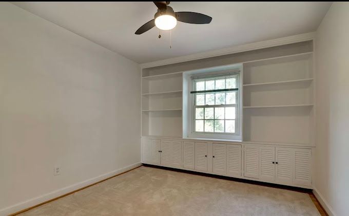 Empty room with beige carpet, white built-in shelves around a window, and a ceiling fan.