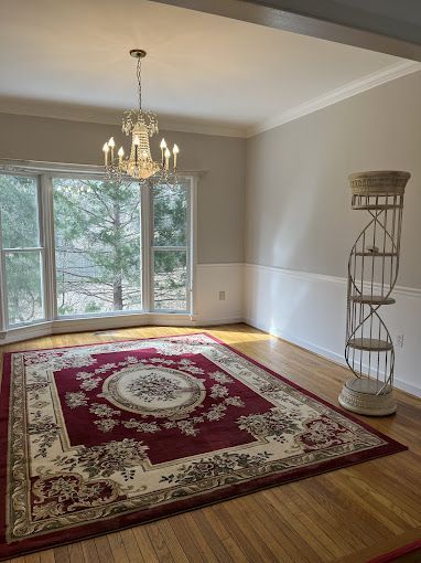 Dining room with red rug, chandelier, spiral shelf, and large window overlooking trees.