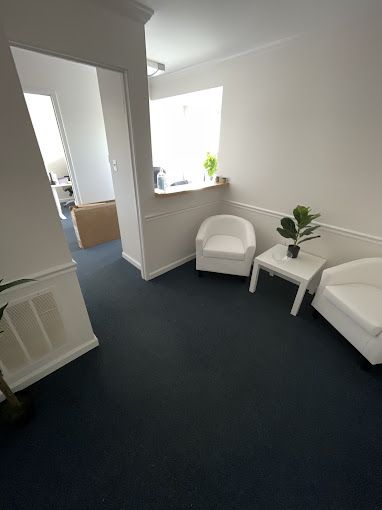 Waiting room with white chairs, small table, and greenery on dark blue carpet.
