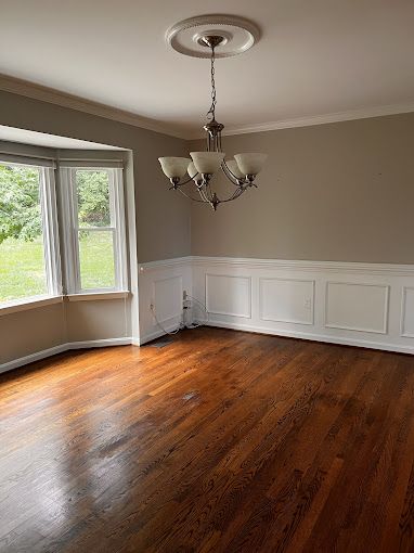 Empty dining room with wood floor, white wainscoting, chandelier, and a bay window.