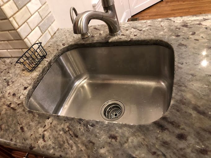Stainless steel kitchen sink in granite countertop. Faucet, drain, and small wire basket are visible.