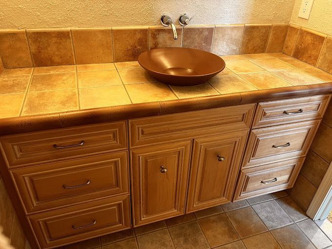 Bathroom vanity with brown vessel sink, light wood cabinets, and tan tile countertop.