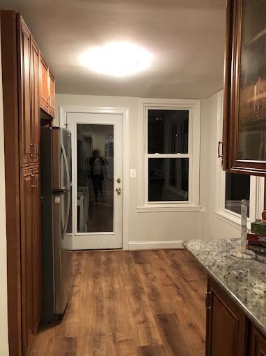 Kitchen interior: wooden cabinets, stainless steel refrigerator, door to outside, window, and wood-look flooring.