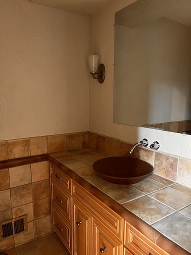 Bathroom with a brown vessel sink on a tiled countertop above wooden cabinets, with a wall mirror and a sconce.