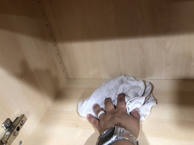 Hand wiping inside of a light-colored wooden cabinet with a white cloth.