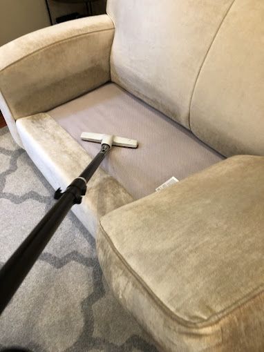 A person vacuuming a light beige sofa with a long black vacuum cleaner wand.