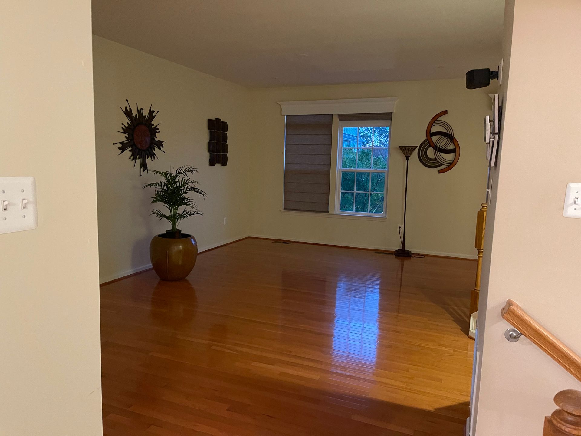 Empty, wood-floored room with a potted plant, sunburst decor, window, and a light-colored wall.