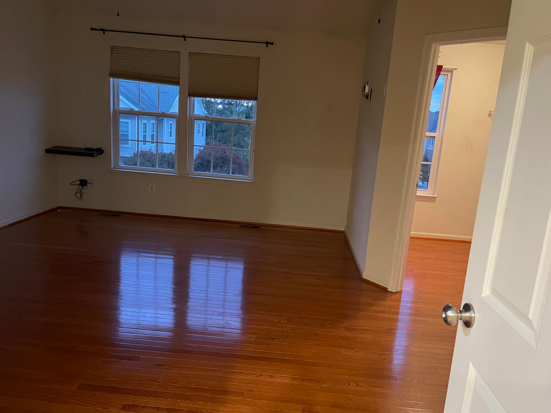 Empty room with hardwood floors, a window with closed blinds, and an open doorway to another room.