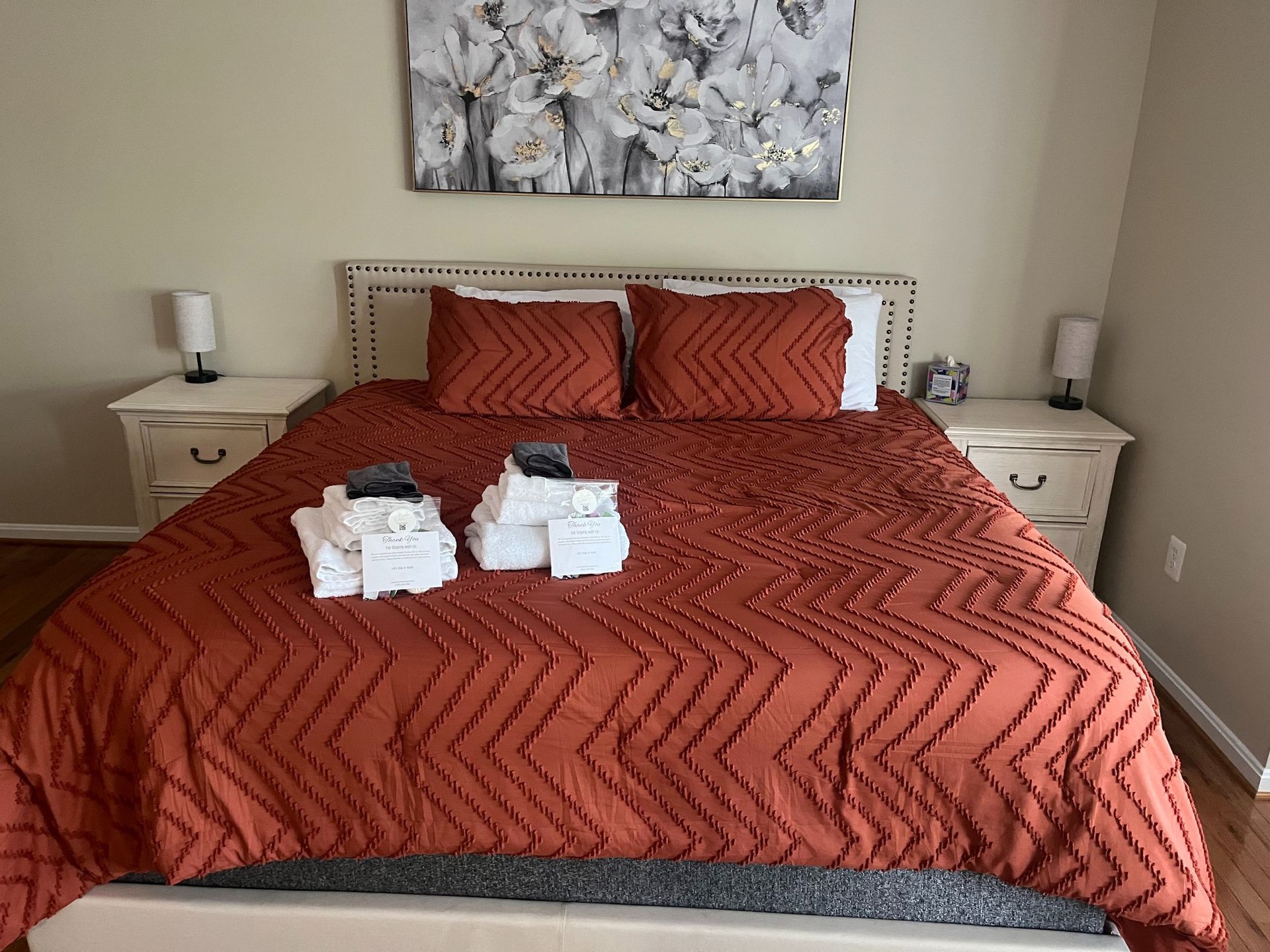 Bedroom with a bed covered in a rust-colored quilt, flanked by matching nightstands, and a floral painting on the wall.
