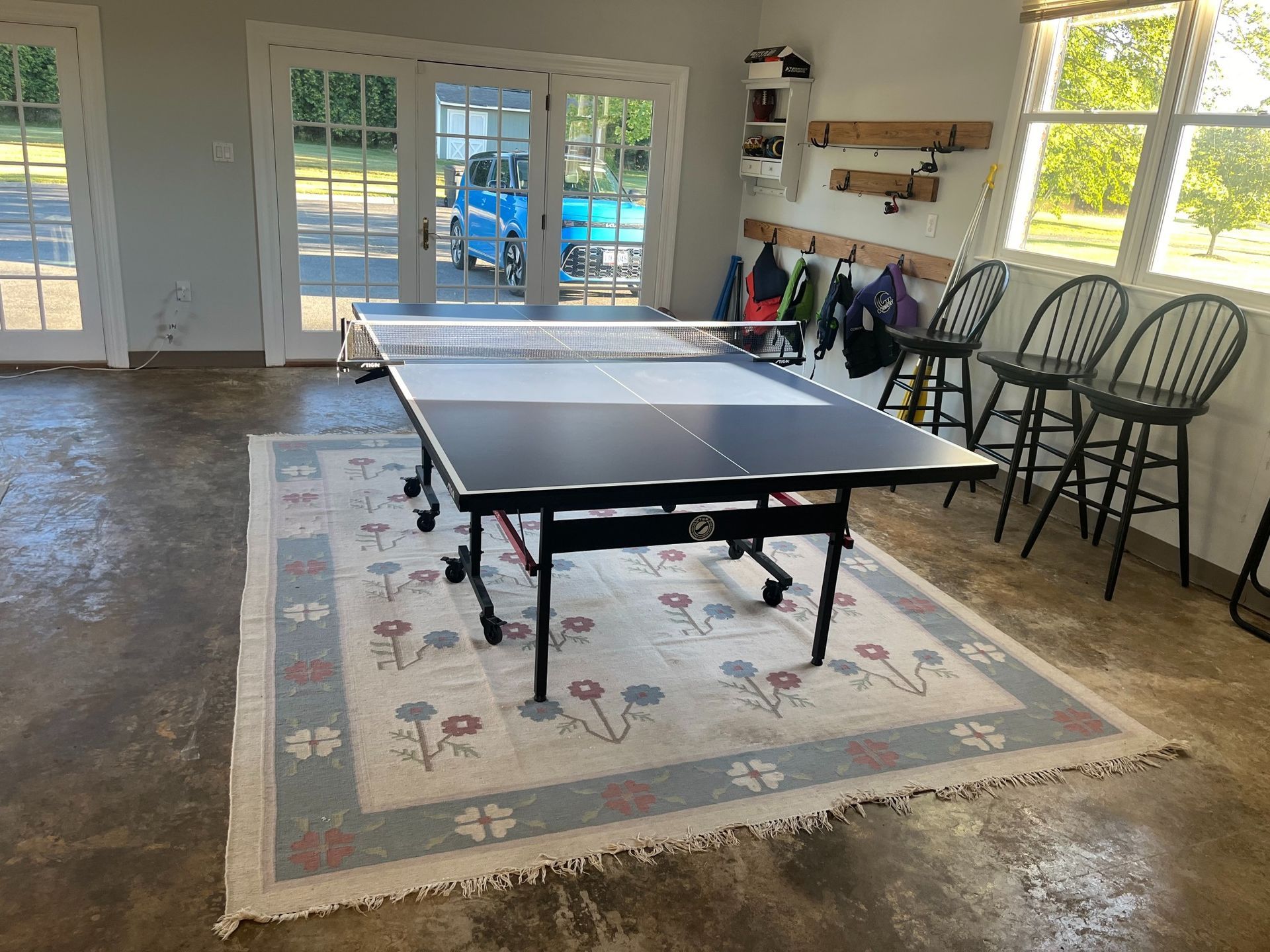 Ping pong table on a floral rug in a garage, with chairs, windows, and french doors to the outside.