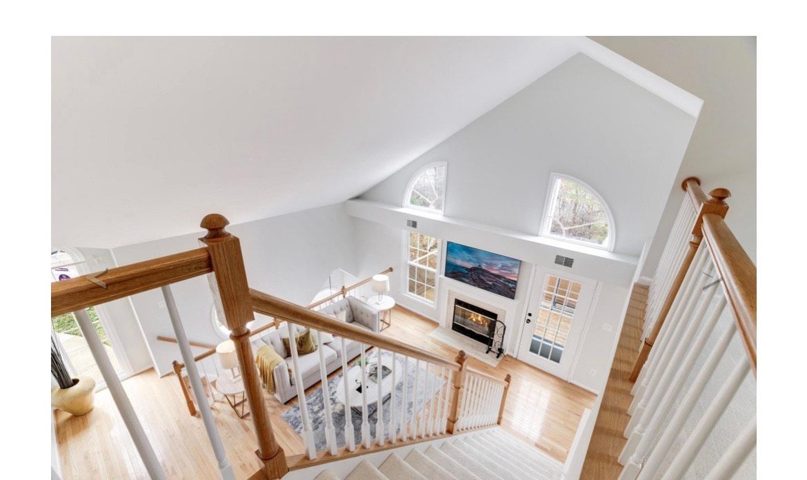 View from a staircase overlooking a bright living room with high ceilings, fireplace, and windows.