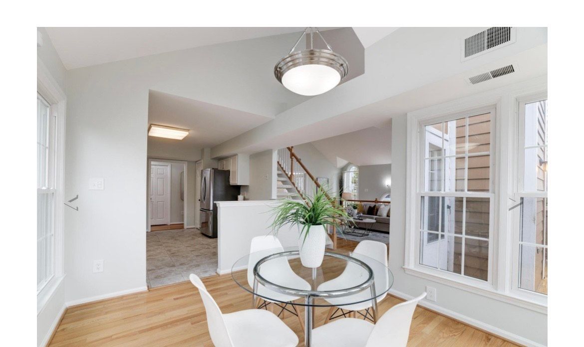 Dining area with glass table and white chairs, light wood floors, and view to kitchen and living room.