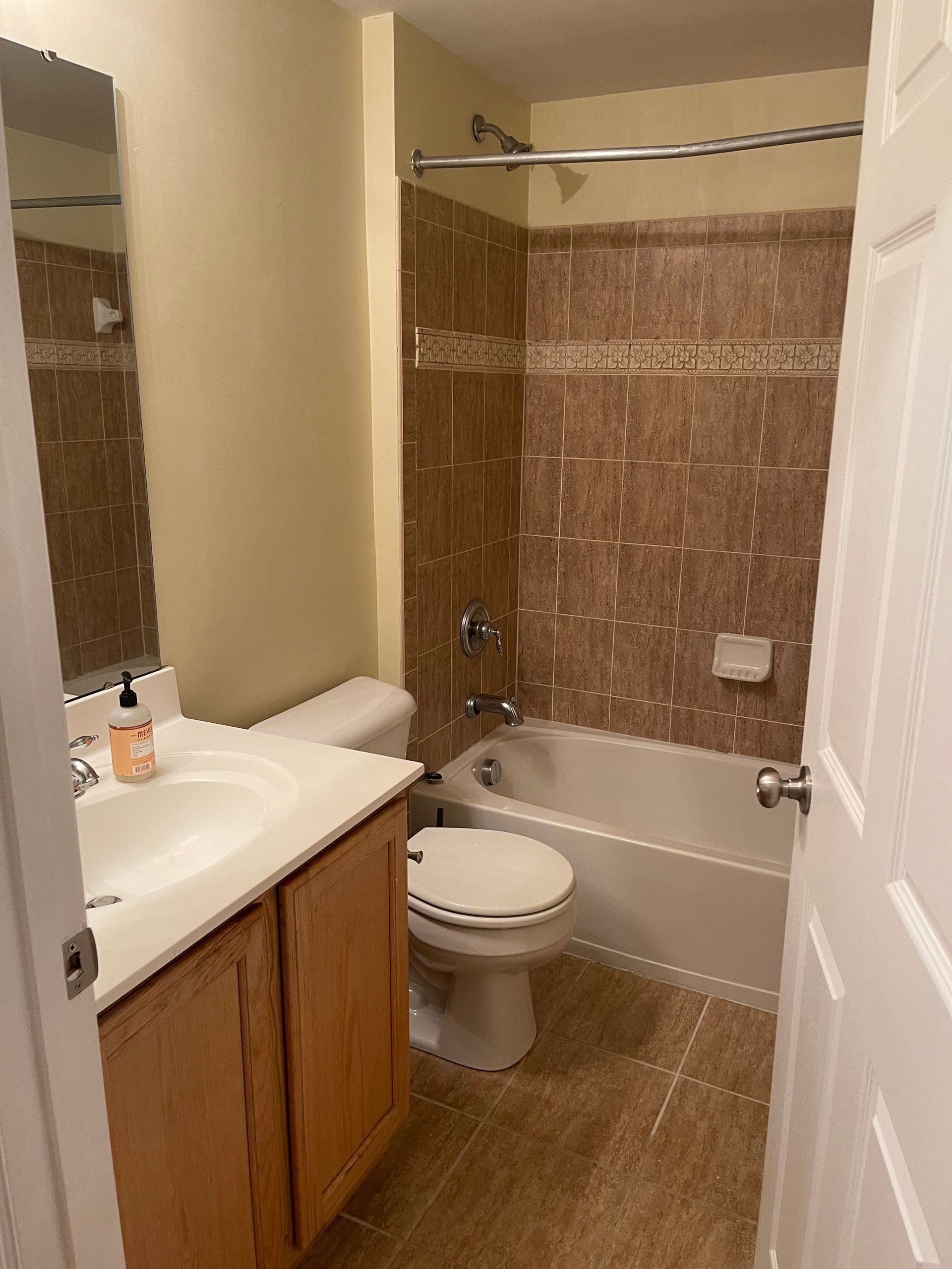 Bathroom with tan walls, brown tile, white tub, and wooden vanity.