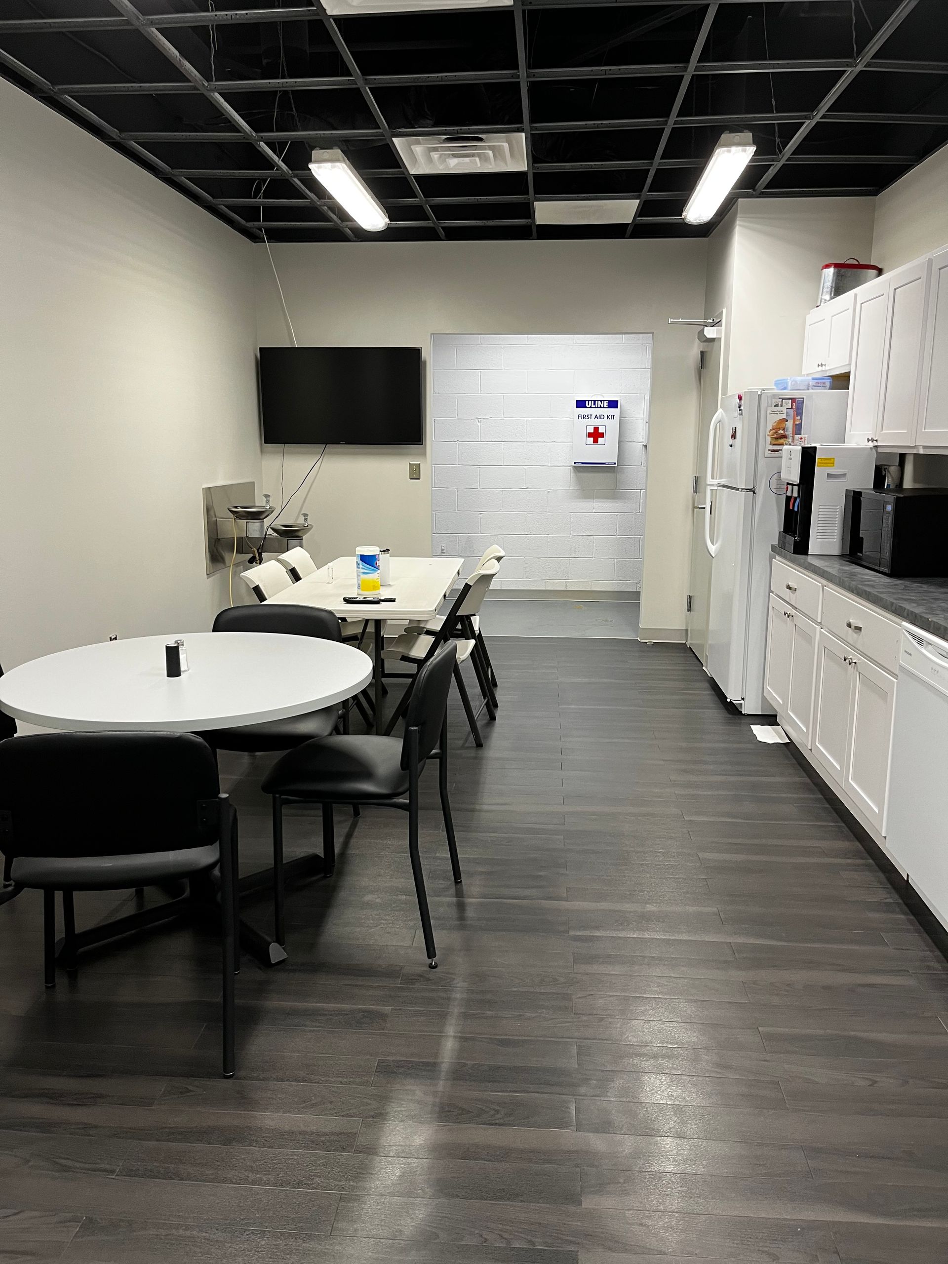 Interior view of an office break room with tables, chairs, kitchen appliances, and access to a hallway.