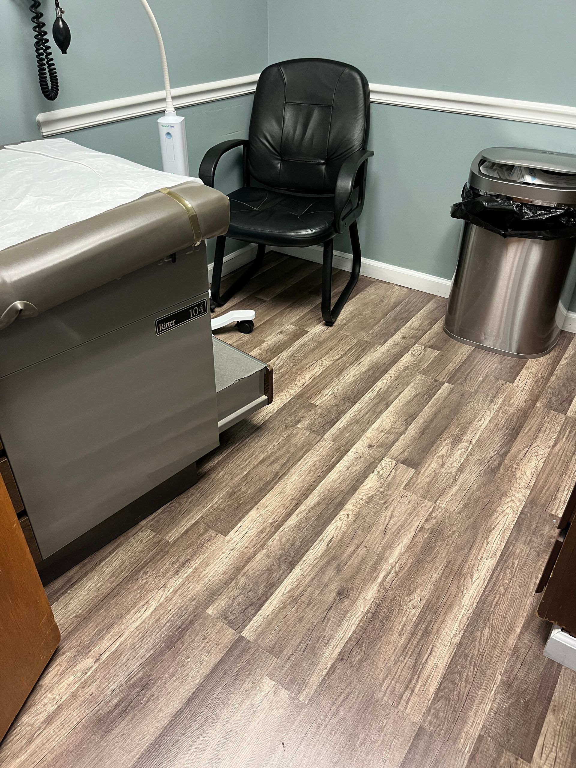 Doctor's office exam room with a chair, exam table, trash can, and medical equipment. Brown and blue tones.