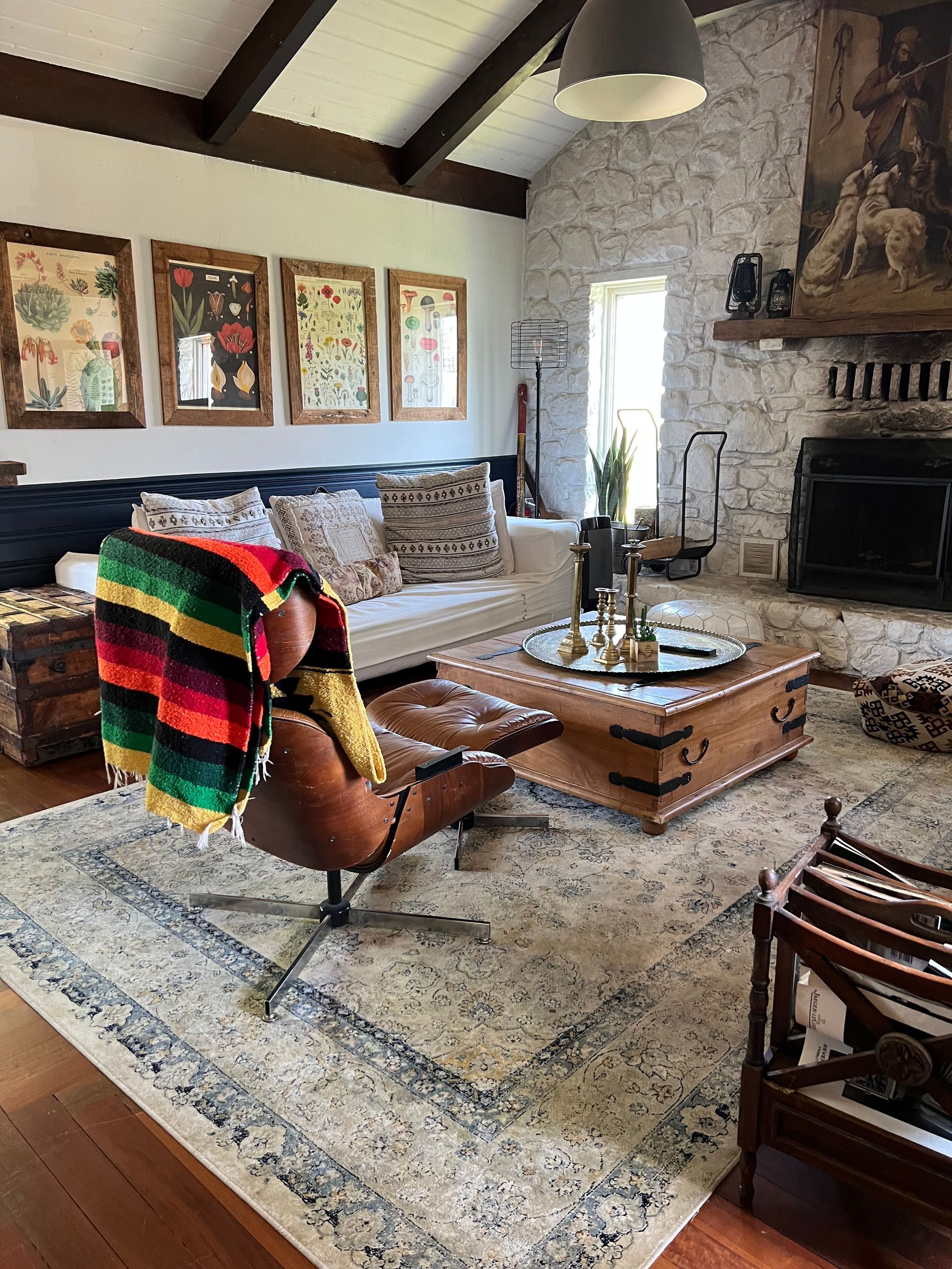 Cozy living room with leather chair, fireplace, white stone wall, and wooden beams.  Rug, coffee table, and framed art.