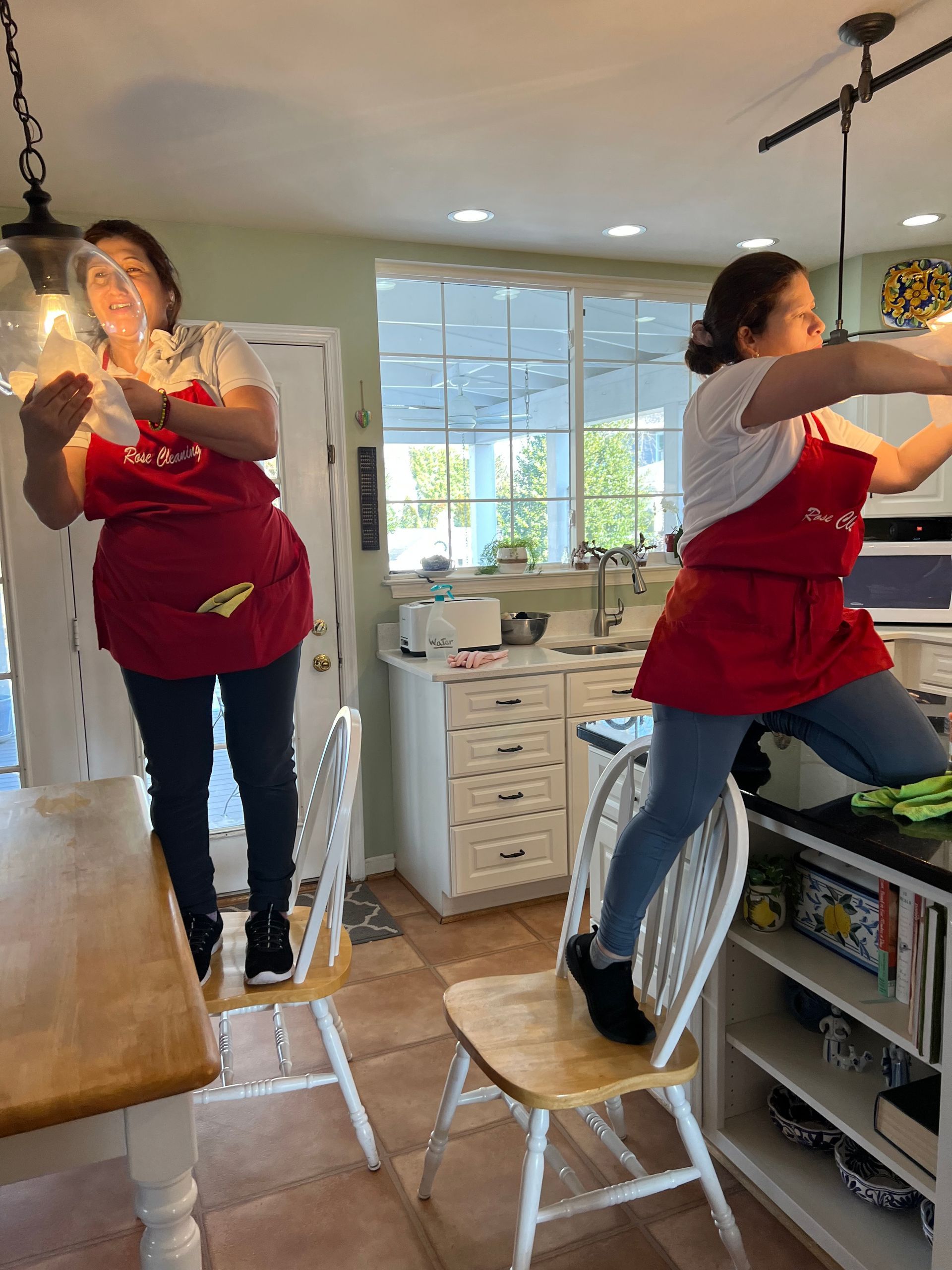 Woman standing on chair cleaning kitchen. Wearing apron, white shirt, black leggings.