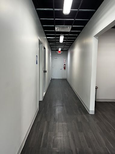 Long hallway with white walls, black ceiling tiles, dark wood-look floor, and a closed door at the end.