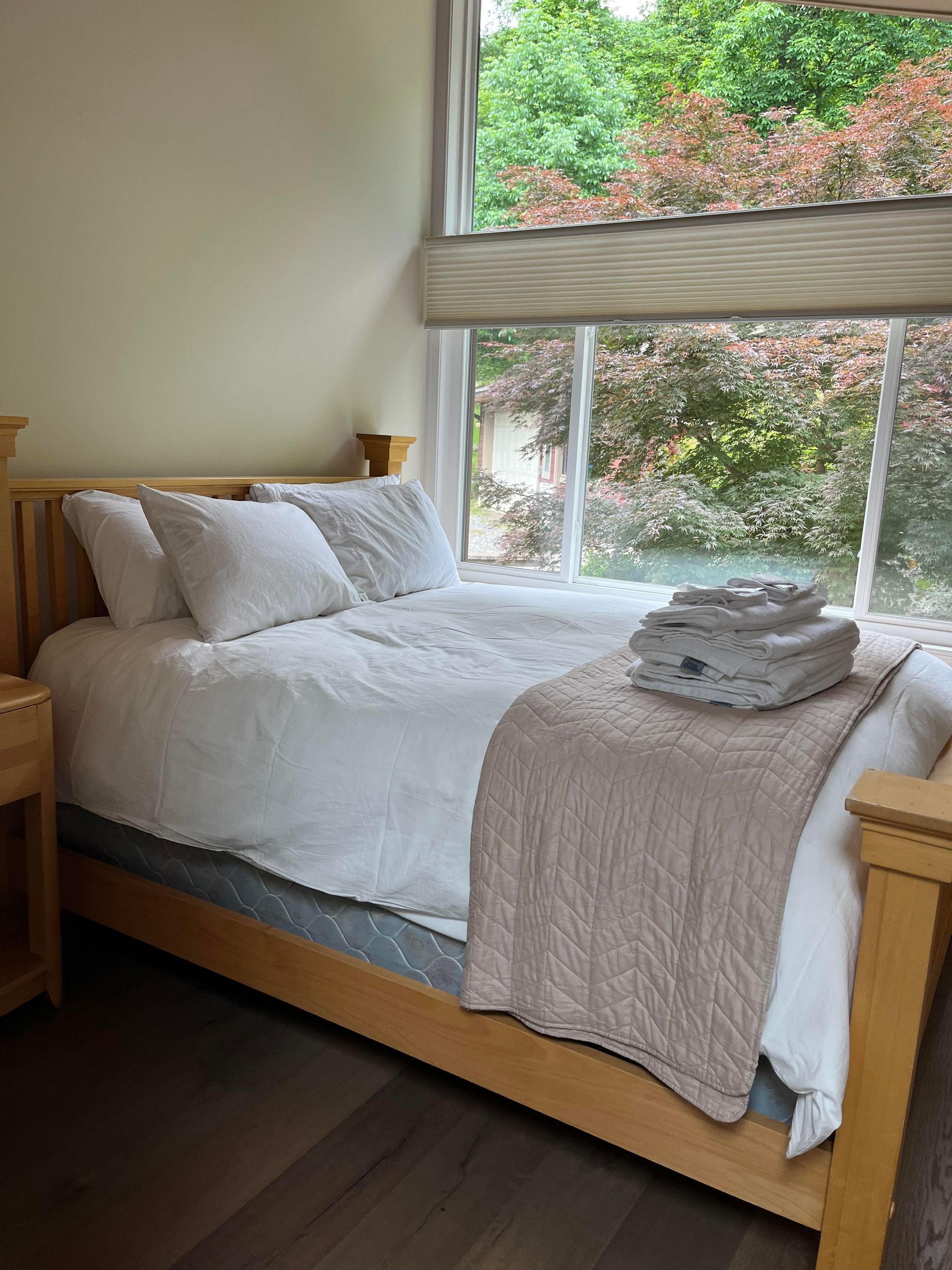 Bedroom with light wood bed frame, white bedding, and window overlooking greenery.