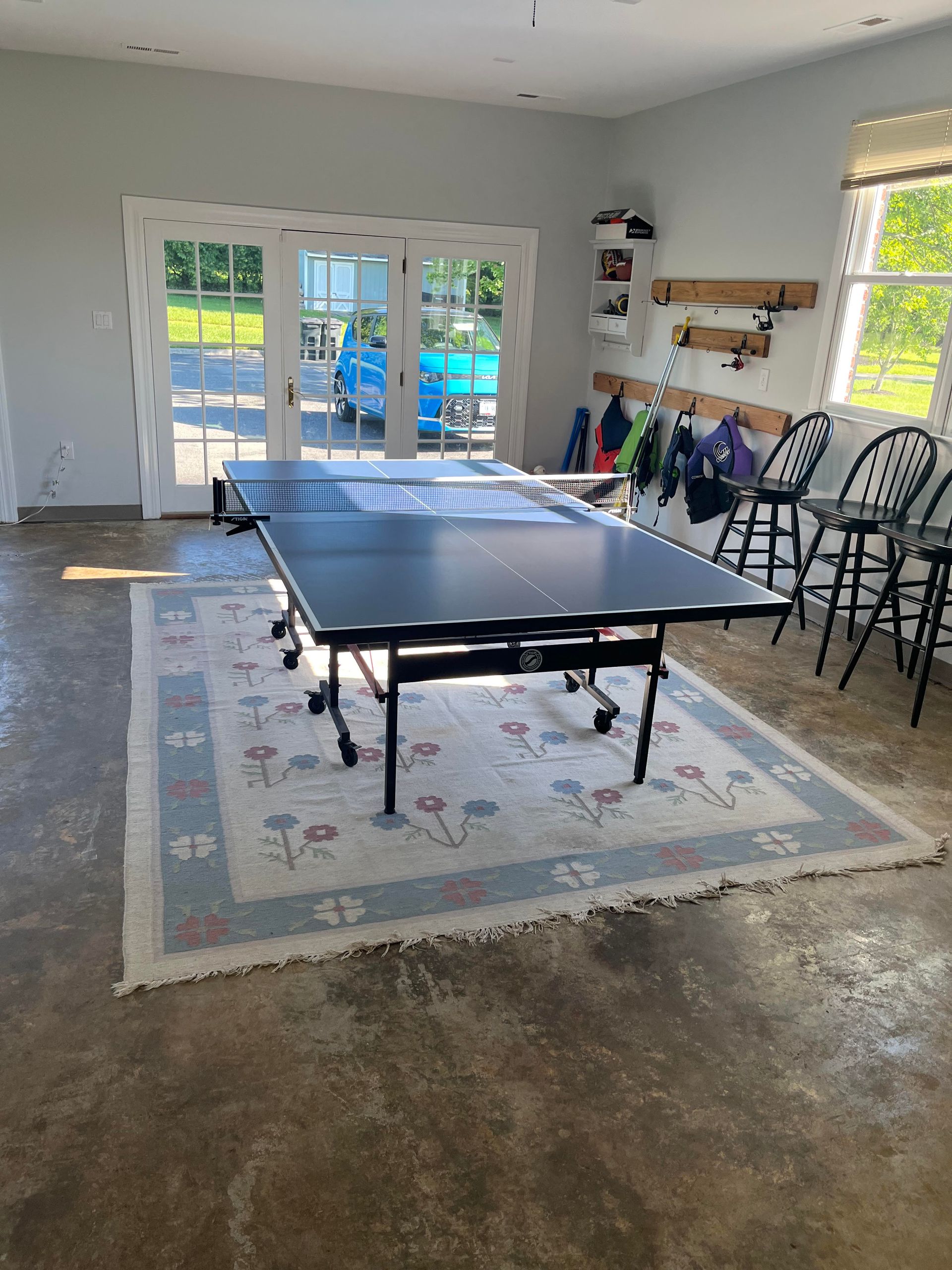 Ping pong table on a floral rug in a garage. French doors, bar stools, and gear on a wooden rack.