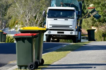 Green and yellow trash bins on a residential street as a white garbage truck approaches to collect them.