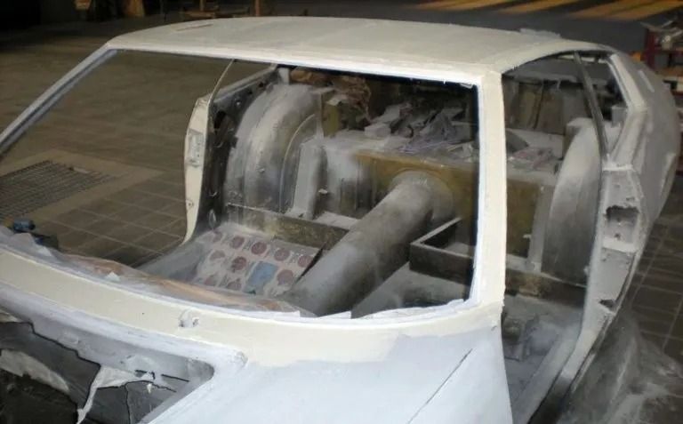 Telaio dell'auto parzialmente assemblato in officina, primer bianco sulla carrozzeria, componenti interni visibili.