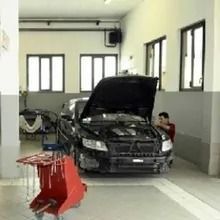 Auto con il cofano aperto in un garage, meccanico al lavoro sul motore. Carrello portautensili rosso in primo piano, finestre sullo sfondo.