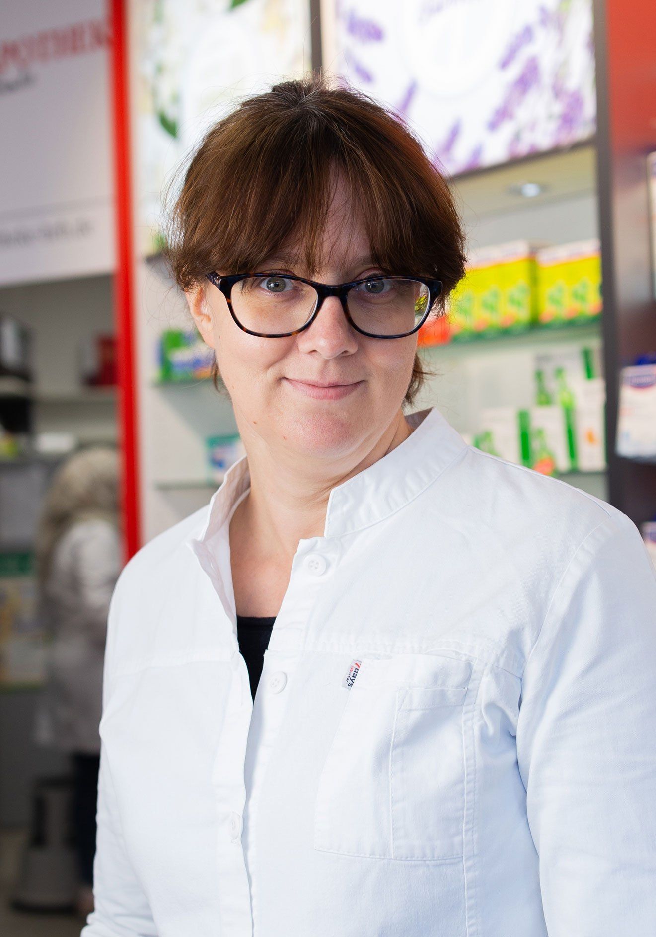 Eine Frau mit Brille und weißem Kittel steht in einer Apotheke.