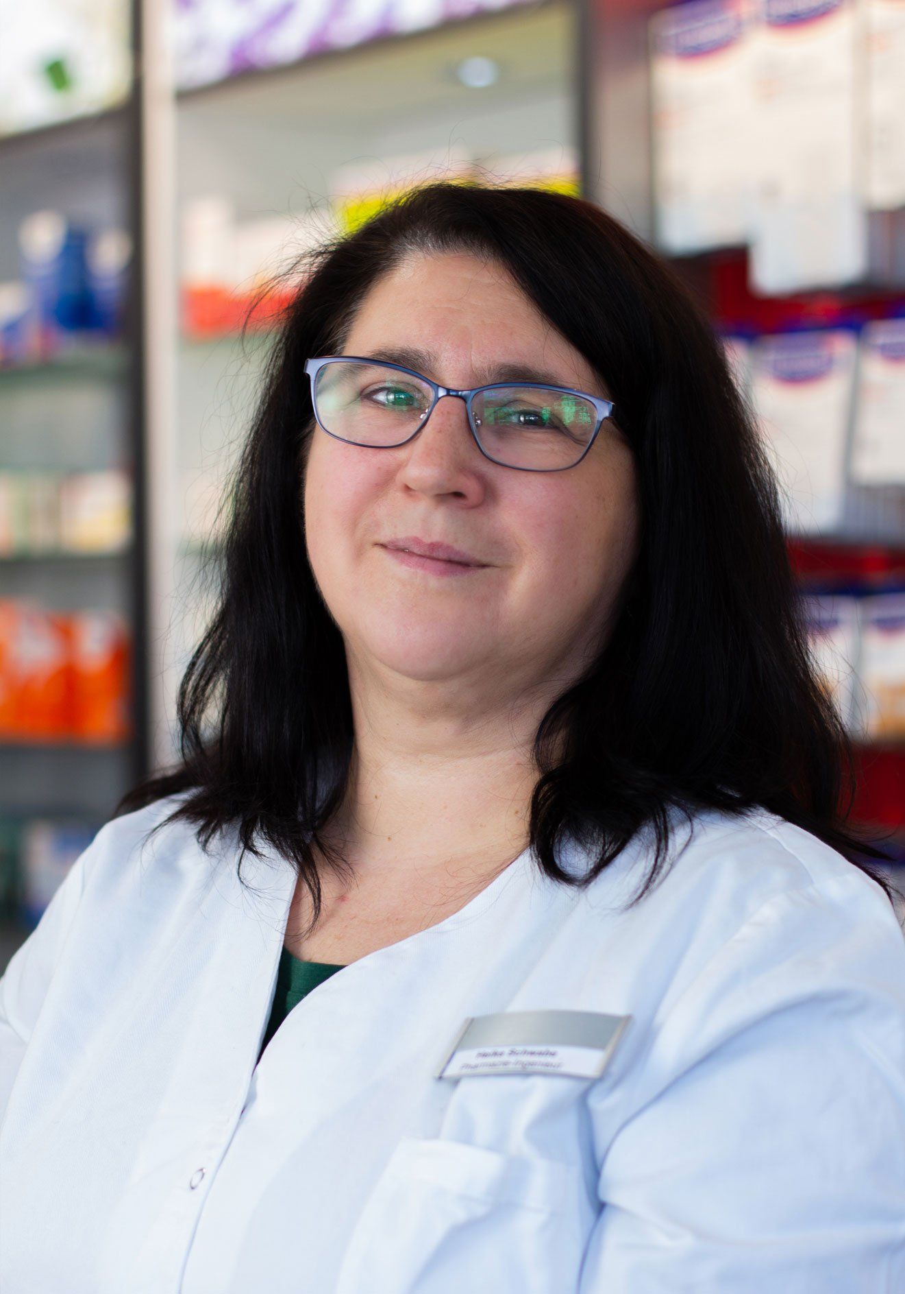 Eine Frau mit Brille und weißem Kittel steht vor einem Regal in einer Apotheke.