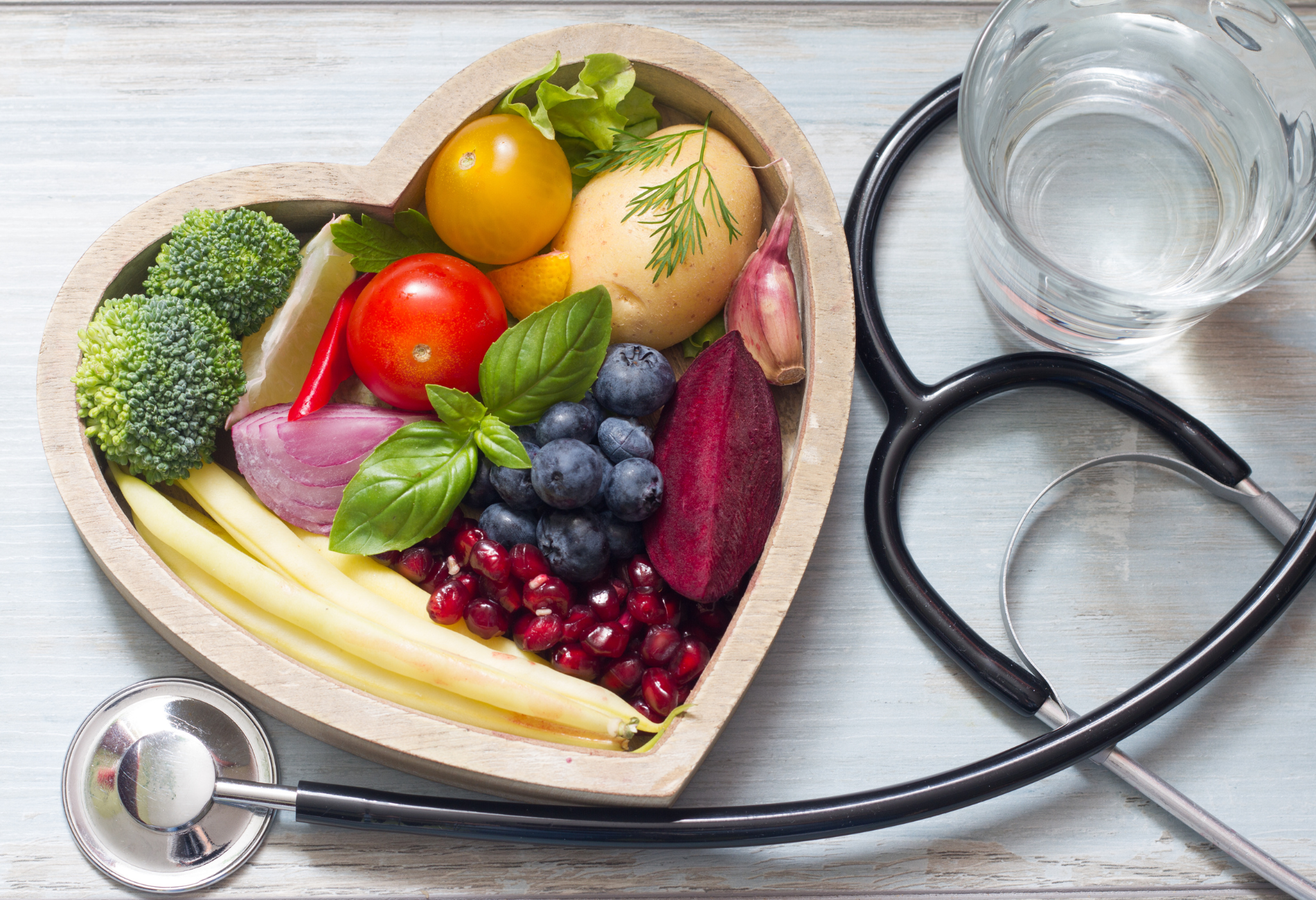a heart shaped bowl filled with fruits and vegetables next to a stethoscope and a glass of water .