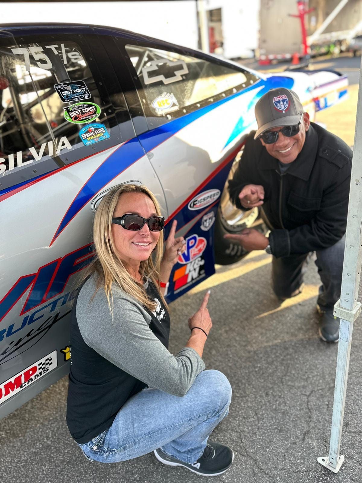 Two people point at a race car. Woman kneels near the car, man kneels beside her. Car is blue, white, and gray. Outdoors.