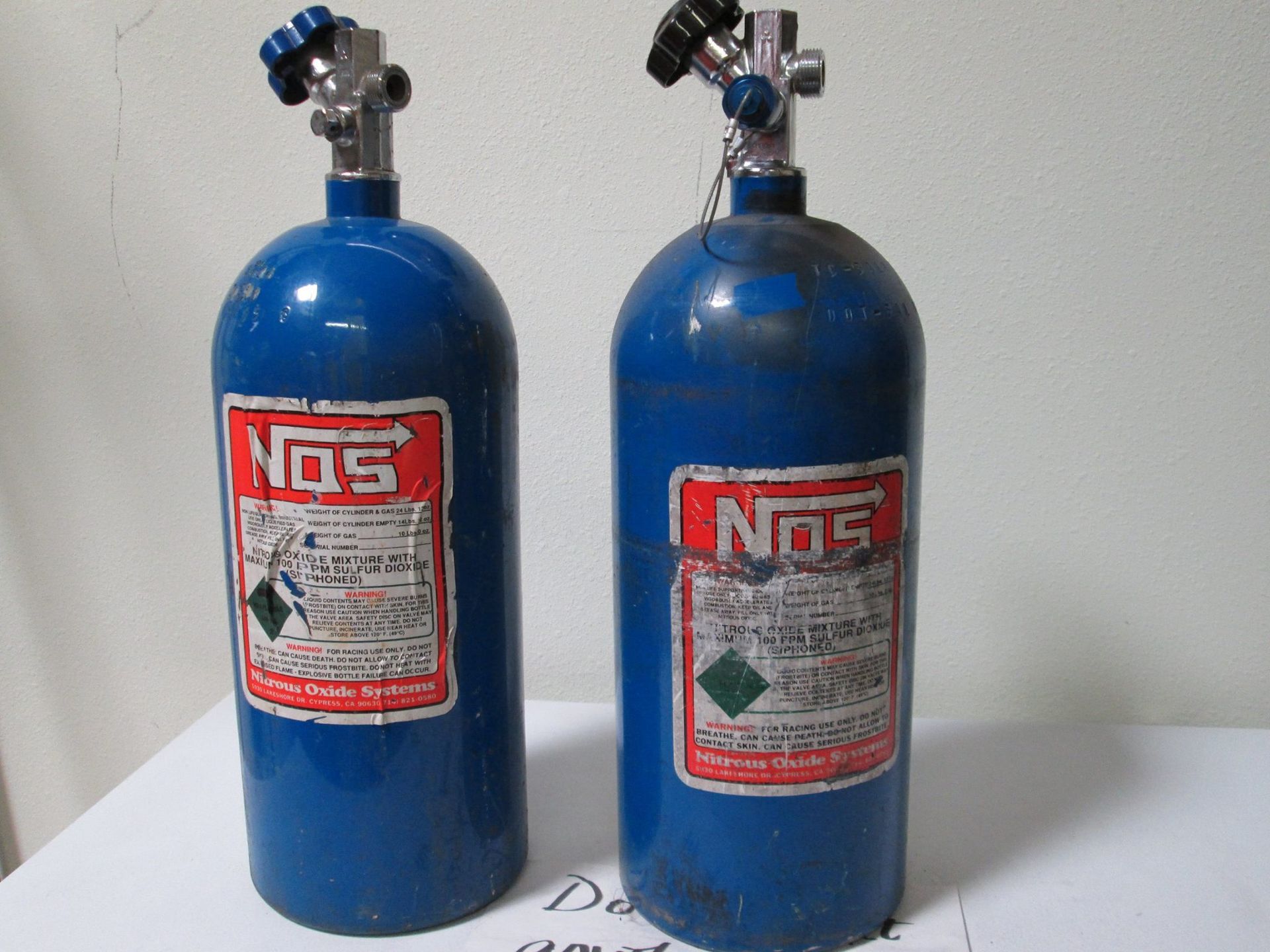Two blue NOS nitrous oxide tanks with red and white labels, set against a white wall.