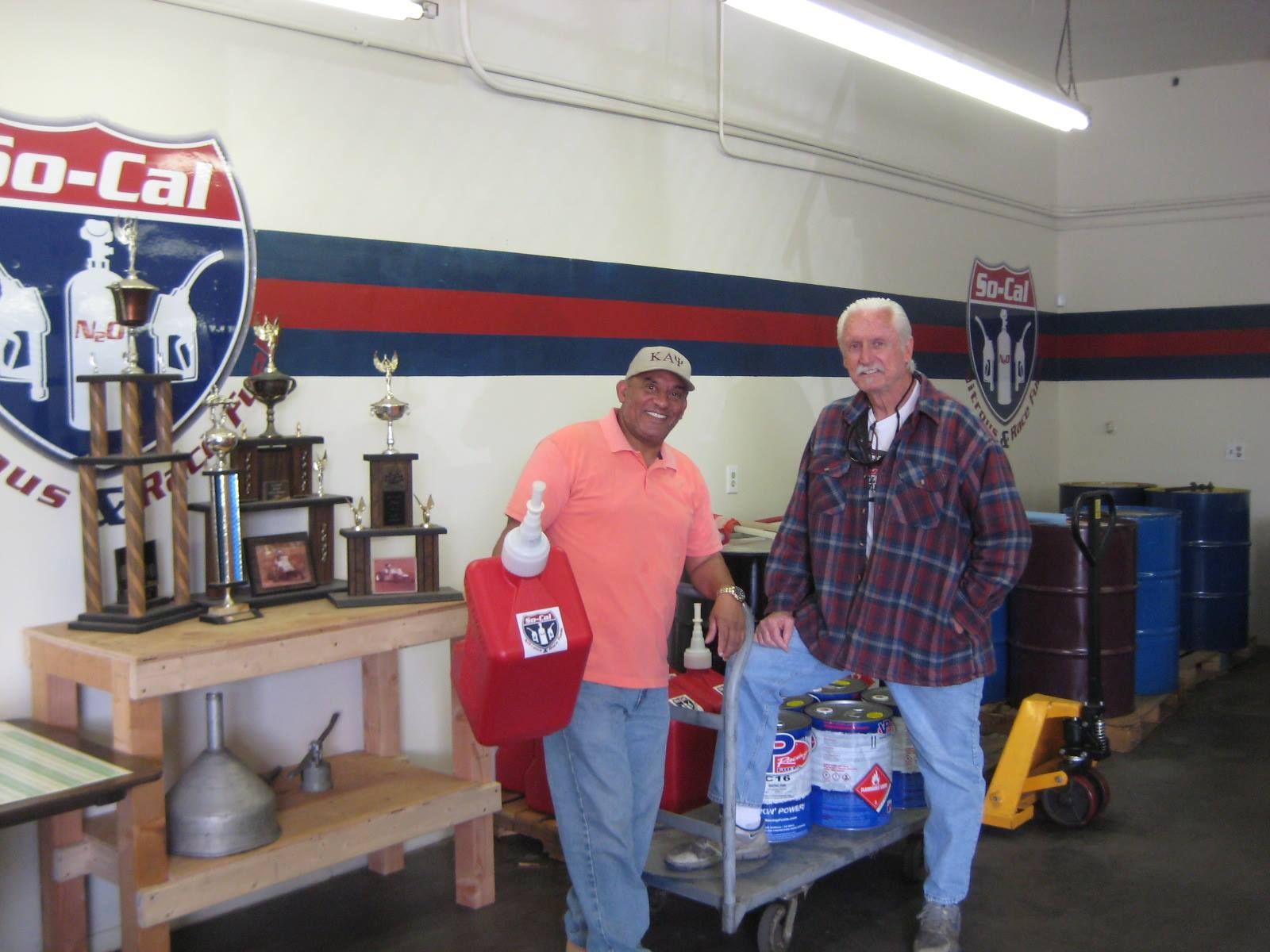 Two men in a shop with trophies, one holding a red gas can. Shop features red/blue stripes.