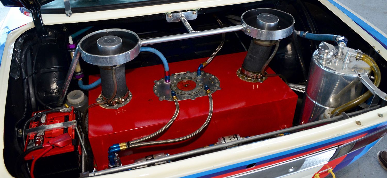 Red fuel tank with two intake tubes, a silver canister, and blue hoses in a car's trunk.