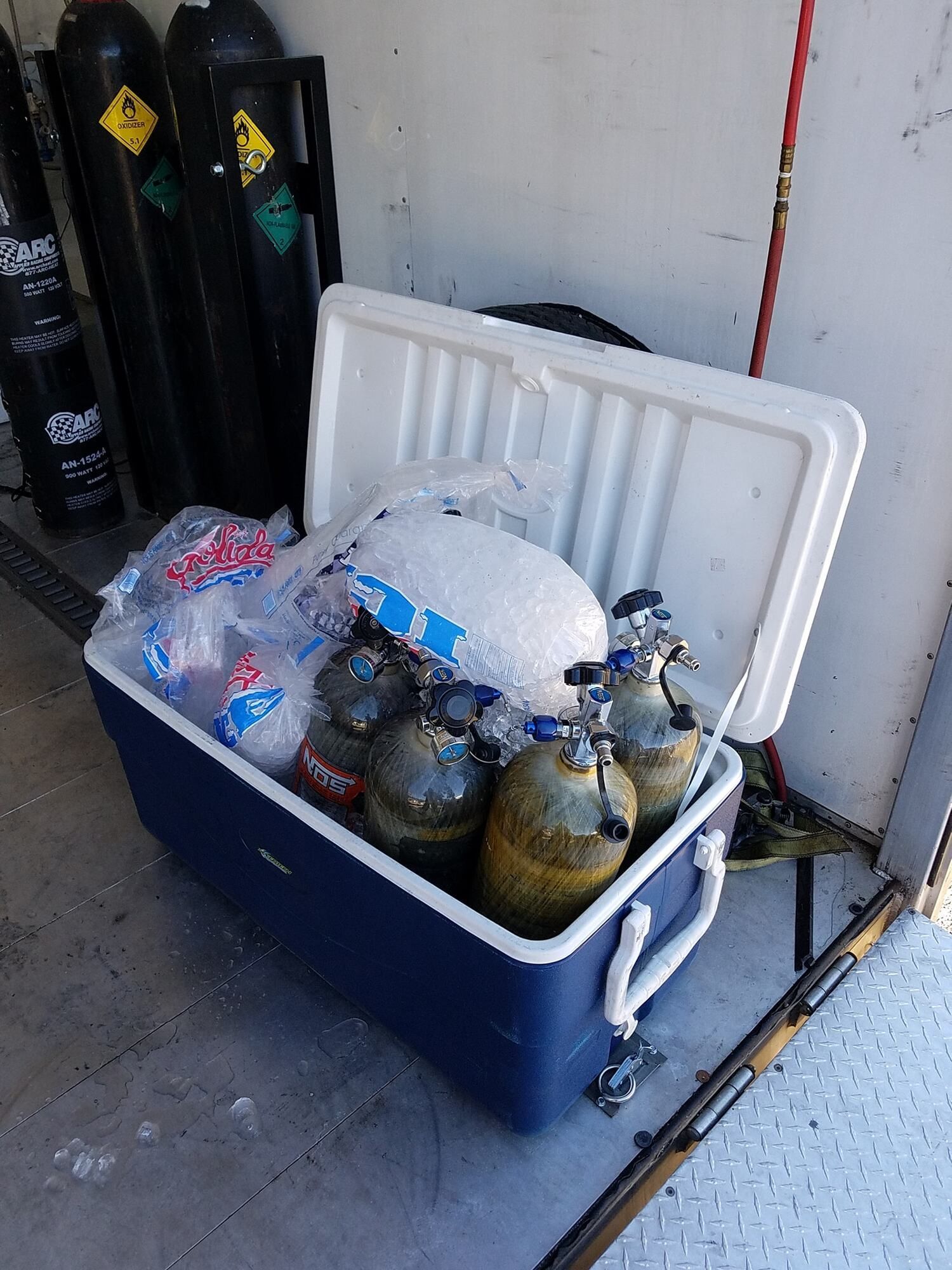 Blue cooler with scuba tanks and ice next to compressed gas cylinders.