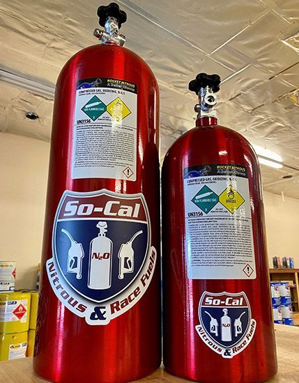 Two red So-Cal Nitrous & Race Fuels tanks of different sizes with safety labels in an indoor setting.
