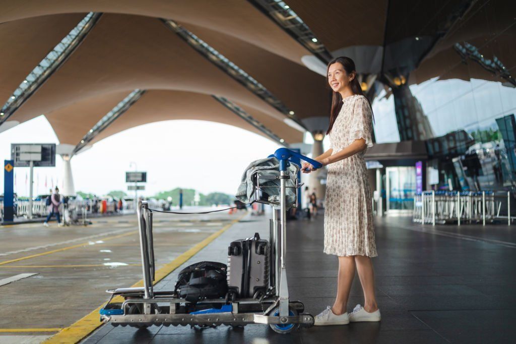 A smiling middle-aged lady is holding a trolley carrying her luggage waits outside the airport.