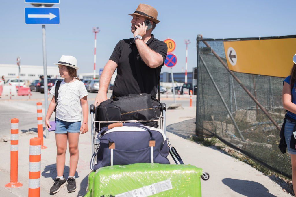 A man on the phone holds his trolley full of luggage while his daughter stands beside him.