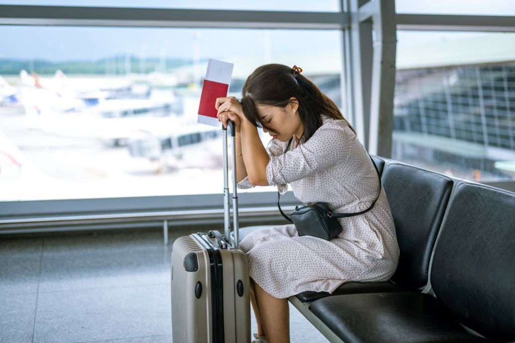 A stressed Asian woman sits and leans on her suitcase in frustration.