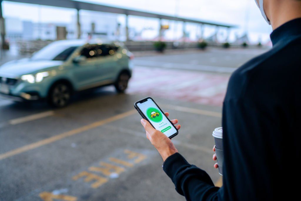 A person holding a mobile phone with a rideshare app on one hand and a coffee cup on the other.