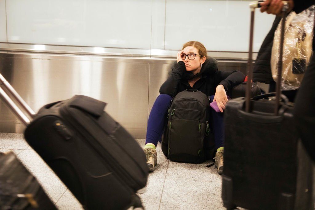 A frustrated Caucasian woman sits on the floor with her backpack surrounded by suitcases.
