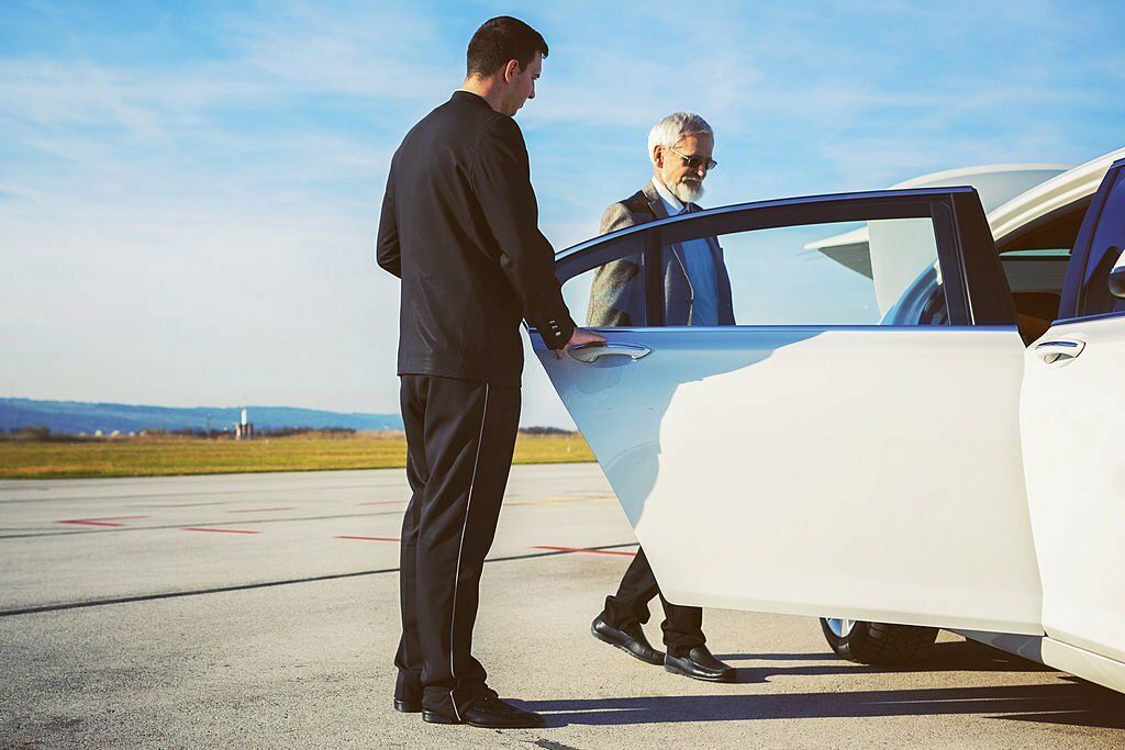 A limo chauffeur is helping a senior adult man into a limousine at the airport.