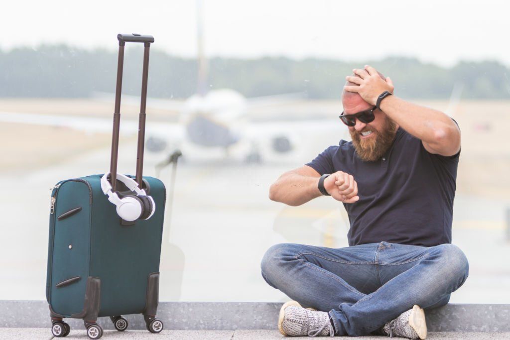 A frustrated man is sitting beside his luggage and looks at his watch in horror and frustration.